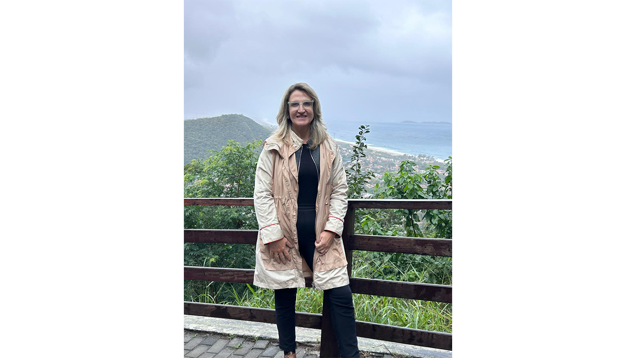 Em um mirante, uma mulher de óculos, vestindo casaco bege e roupa preta, está em pé diante da câmera. Ao fundo, aparecem vegetação, morros, faixa litorânea e parte da área urbana.