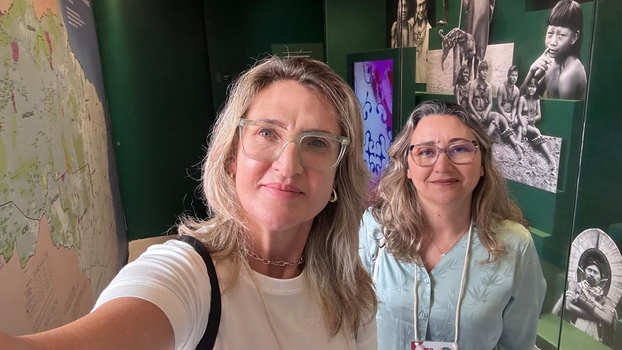 Em uma exposição, duas mulheres de óculos posam para uma foto estilo selfie; uma veste blusa branca de mangas curtas e a outra, blusa azul-clara de mangas compridas com crachá de visitante. Ao fundo, há mapas, fotografias em preto e branco e painéis de temática cultural, com retratos de povos indígenas e um fundo verde.
