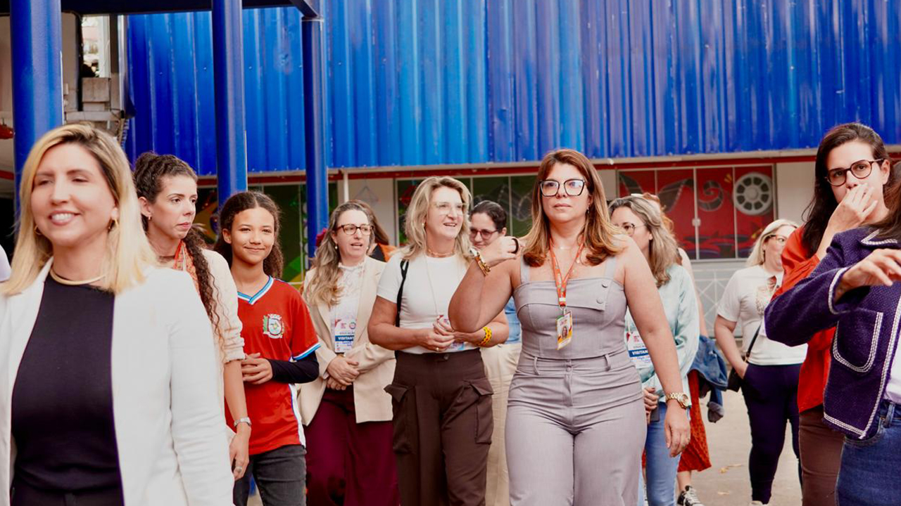 Área externa de uma escola, com ao menos 12 pessoas caminhando em grupo, todas mulheres. Elas usam roupas casuais e crachás, e uma estudante veste uma camiseta vermelha e calça jeans. Ao fundo, há uma estrutura azul e a fachada escolar.