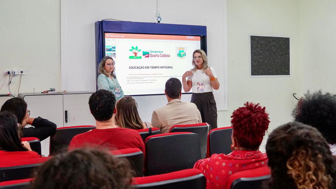 Sala de apresentação de paredes claras e público sentado em cadeiras com estofado vermelho. Duas mulheres estão em pé diante de uma tela de projeção; uma delas gesticula enquanto apresenta o conteúdo “Geoparque Quarta Colônia - Educação em Tempo Integral”.