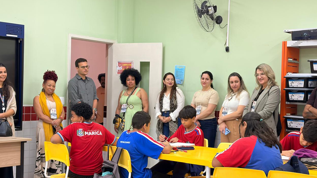 Sala de aula com estudantes sentadas(os) em mesas e cadeiras amarelas, realizando atividades. Um grupo multirracial de oito adultos com crachás observa as(os) alunas(os), que vestem roupas vermelhas e azuis com a inscrição "Prefeitura de Maricá" nas costas, juntamente com o brasão da cidade. Ao fundo, há armários, estantes e paredes verdes. A porta branca da sala está aberta e há um vestilador fixado na parede.