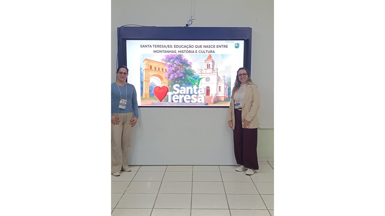 Uma dupla de mulheres de óculos e crachás posam cada uma de um lado de uma tela digital. A tela exibe o texto “Santa Teresa/ES: Educação que nasce entre montanhas, história e cultura”, acompanhado de ilustrações de pontos locais. Ao fundo, há uma parede clara e o piso é de cerâmica clara.