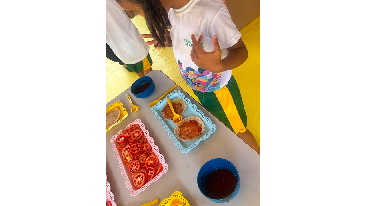 Uma criança vestindo roupas brancas e verdes está em pé, realizando uma atividade de culinária. Sobre a mesa cinza, há bandejas de plástico azul, rosa e amarelo com rodelas de toamte, e mini pizzas com molho de tomate.