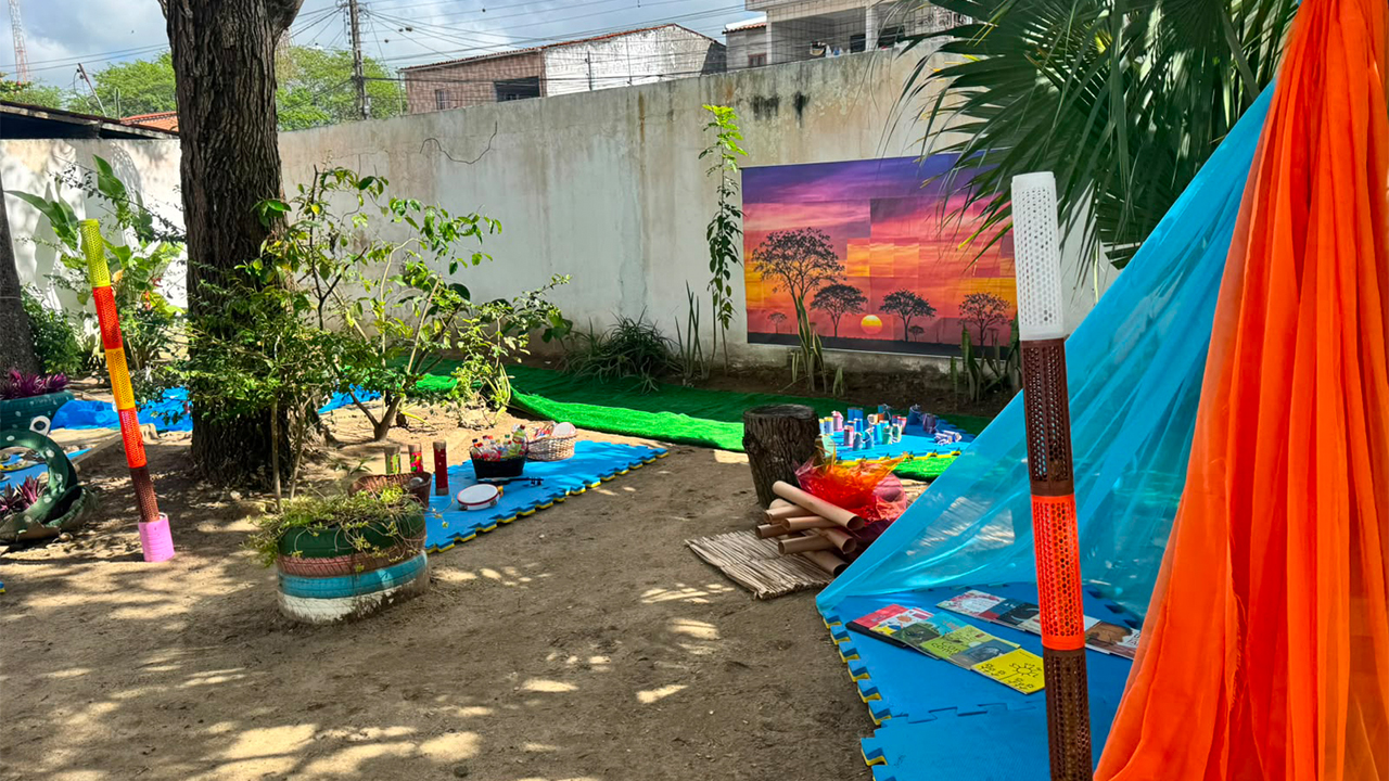Em um quintal decorado, destaca-se, à direita, uma cabana feita com tecidos laranja e azul e longos bastões coloridos fincados no chão. Ao fundo, observa-se um muro branco com um painel pintado representando o pôr do sol, cercado por árvores e tapetes azuis de EVA no chão de areia e terra batida.