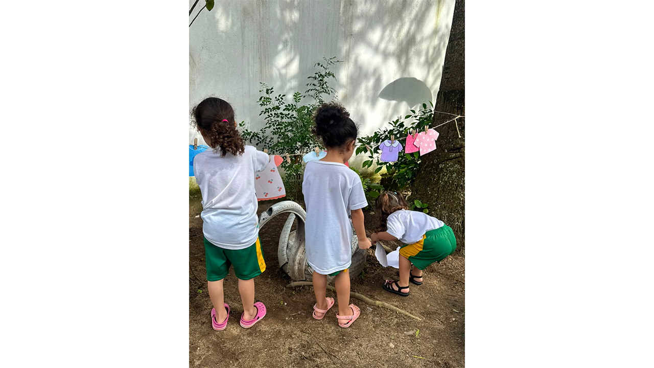Três crianças, de costas, usando roupas semelhantes nas cores branca e verde, interagem próximas a um varal, com roupinhas coloridas de papel presas por pregadores. Uma das cordas do varal está presa no tronco de uma árvore à direita. O espaço tem arbustos verdes e um muro alto e branco ao fundo, além do chão de areia e terra batida.