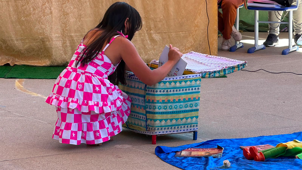 Uma criança está agachada de costas, usando um vestido rosa e branco. Ela manuseia livros dentro de uma caixa decorada, enquanto outras obras e objetos estão dispostos sobre um tecido azul no chão ao seu lado. Ao fundo, aparecem pessoas sentadas e uma mesa coberta por tecido bege.
