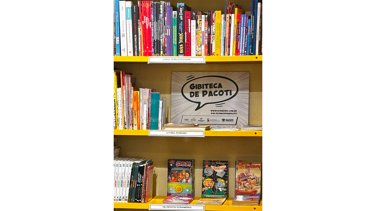 Estante amarela com livros, revistas e histórias em quadrinhos organizados em três prateleiras. Ao centro, uma placa em formato de balão de fala exibe o texto “Gibiteca de Pacoti”.