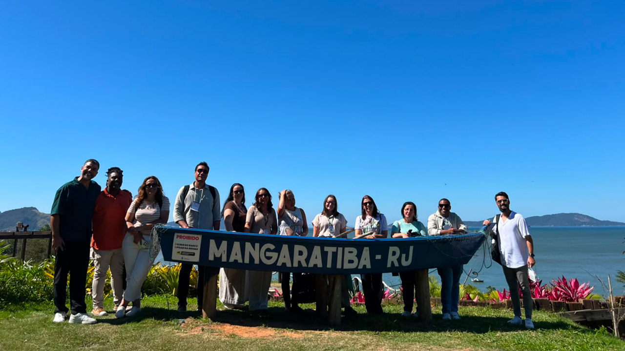 Grupo multiracial de doze pessoas vestindo roupas casuais está em pé, lado a lado, sorrindo. O destaque é uma estrutura de madeira azul, em formato de canoa, à frente deles com o texto "Mangaratiba - RJ", escrito em branco. Ao fundo é possível avistar o mar e montanhas no horizonte, além do céu azul limpo.