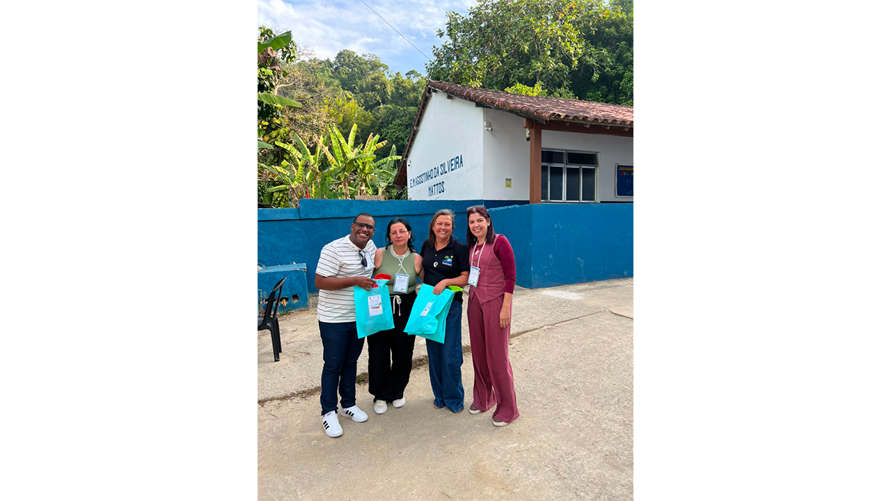 Grupo multirracial formado por um homem e três mulheres veste roupas casuais e posa em pé, lado a lado. O homem e uma das mulheres seguram sacolas azul claro. Ao fundo, a construção branca da escola, cercada por um muro azul e por árvores.