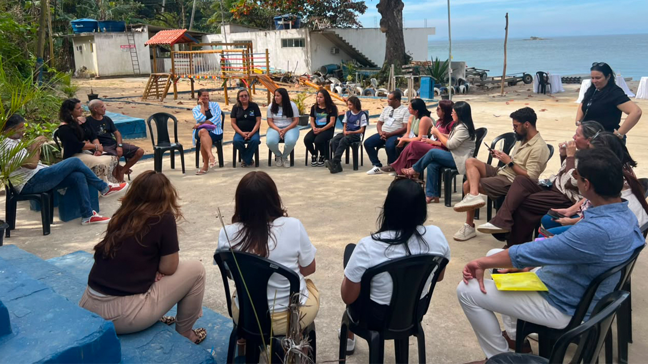 Grupo multirracial de cerca de 20 pessoas está sentado em roda em cadeiras plásticas pretas, conversando entre si. O cenário exibe uma grande árvore ao fundo, um parquinho, construções brancas e o mar ao fundo.