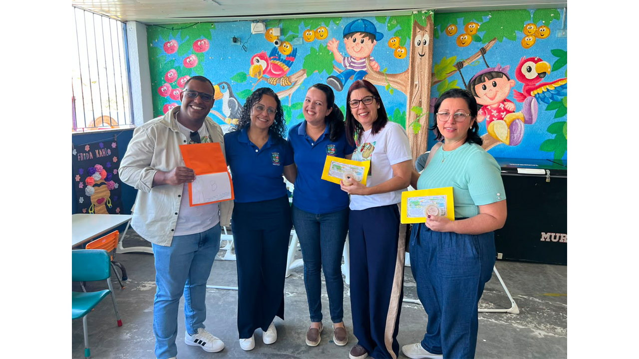 Grupo multirracial de cinco pessoas posa em pé, lado a lado, usando roupas casuais. Duas mulheres e um homem seguram envelopes amarelos e laranjas. Ao fundo, há uma parede com uma pintura infantil colorida de crianças, pássaros, frutas e uma árvore.