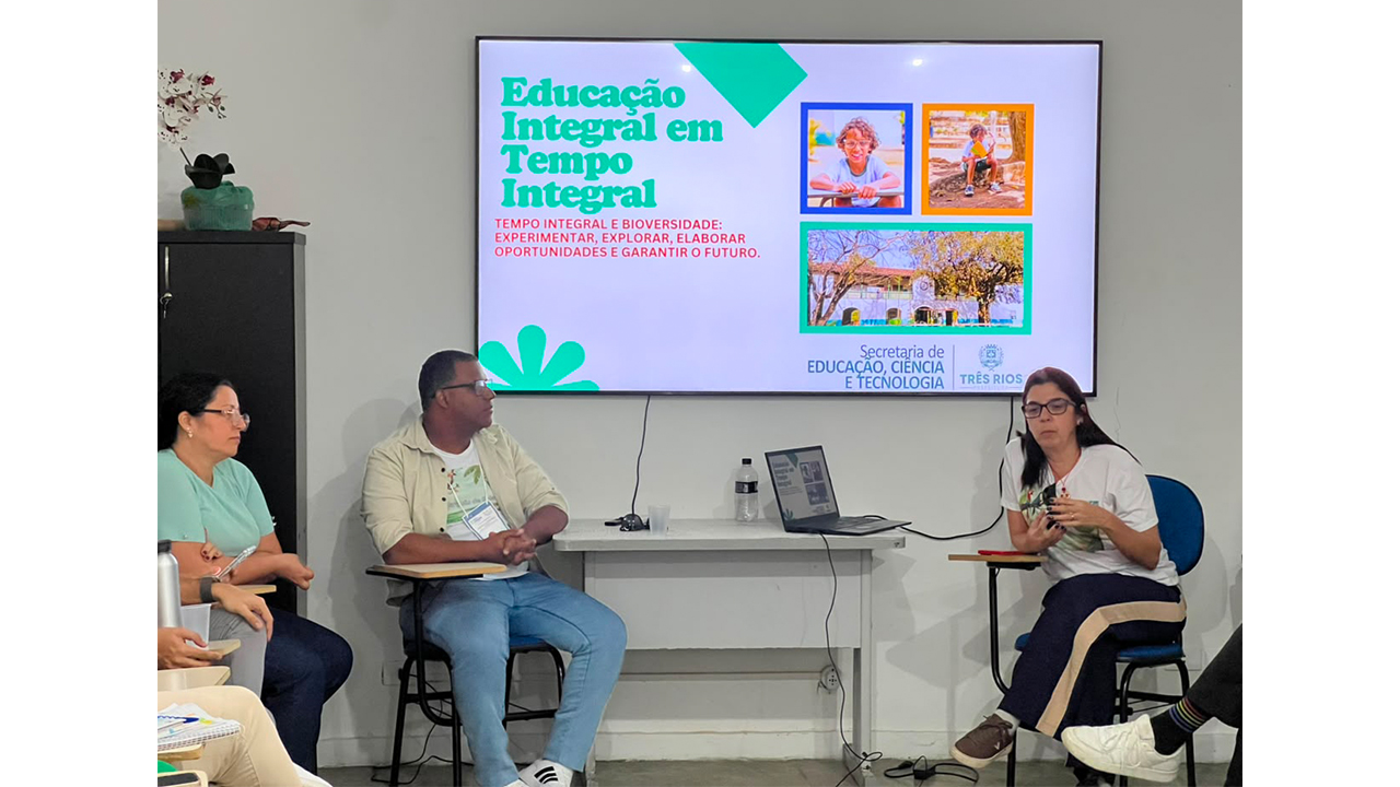 Grupo multirracial formado por três adultos - duas mulheres e um homem - está sentado à frente de uma sala, fazendo uma apresentação. A mulher à direita gesticula enquanto fala, e os outros dois a observam. O destaque é o telão exibindo o título "Educação Integral em Tempo Integral" e fotos de crianças. A sala tem paredes claras e conta uma pequena mesa com um notebook e um armário escuro à esquerda.