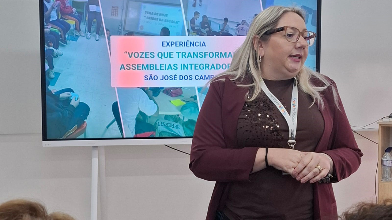 Sala de apresentação com uma mulher de crachá em pé diante do público. Ela veste blusa marrom e um casaco vinho. Ao fundo, uma tela exibe o texto "Experiência 'Vozes que transformam: assembleias integradoras' – São José dos Campos", acompanhada de imagens de atividades escolares com filtro azulado. À frente da mulher, é possível perceber cadeiras de auditório com estofado azul.