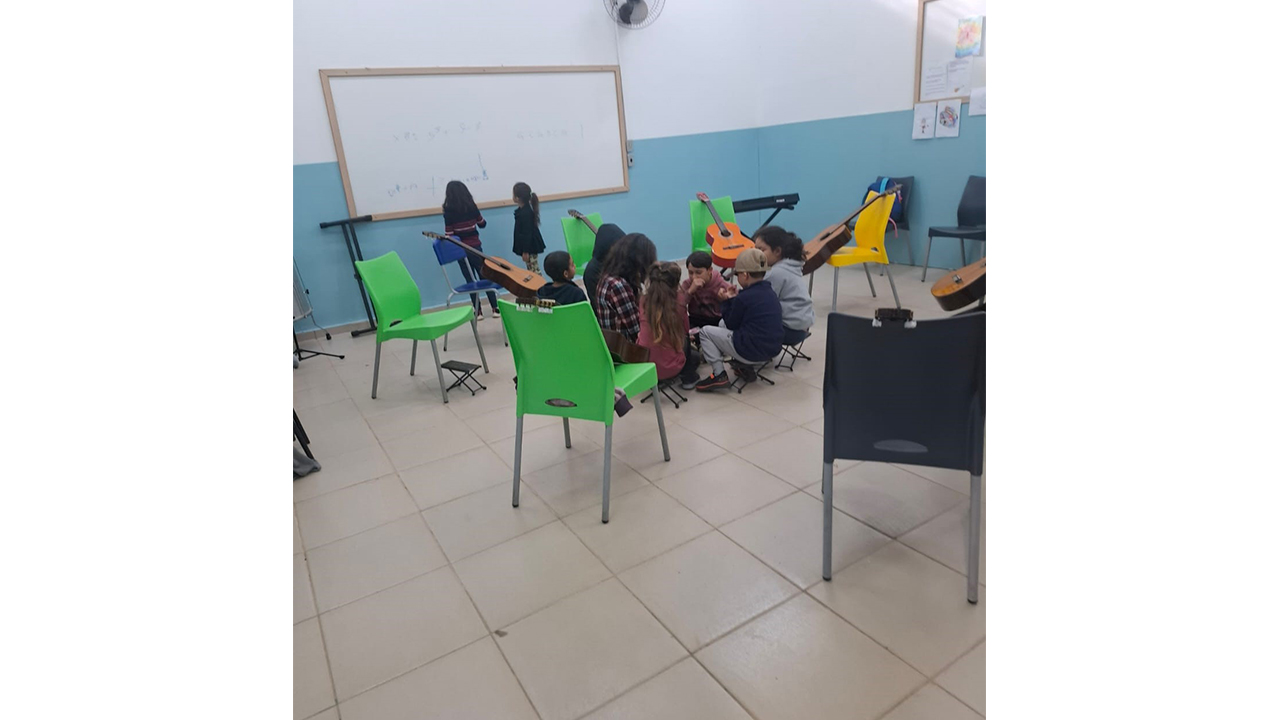 Sala de música com crianças reunidas em pequenos grupos entre cadeiras verdes e cinzas. Violões estão apoiados nas cadeiras e nas laterais, e duas crianças escrevem no quadro branco. Ao fundo, há teclado preto. A sala tem piso claro e as paredes são parcialmente brancas com a metade inferior pintada de azul. Acima, fora do enquadramento, é possível avistar janelas.