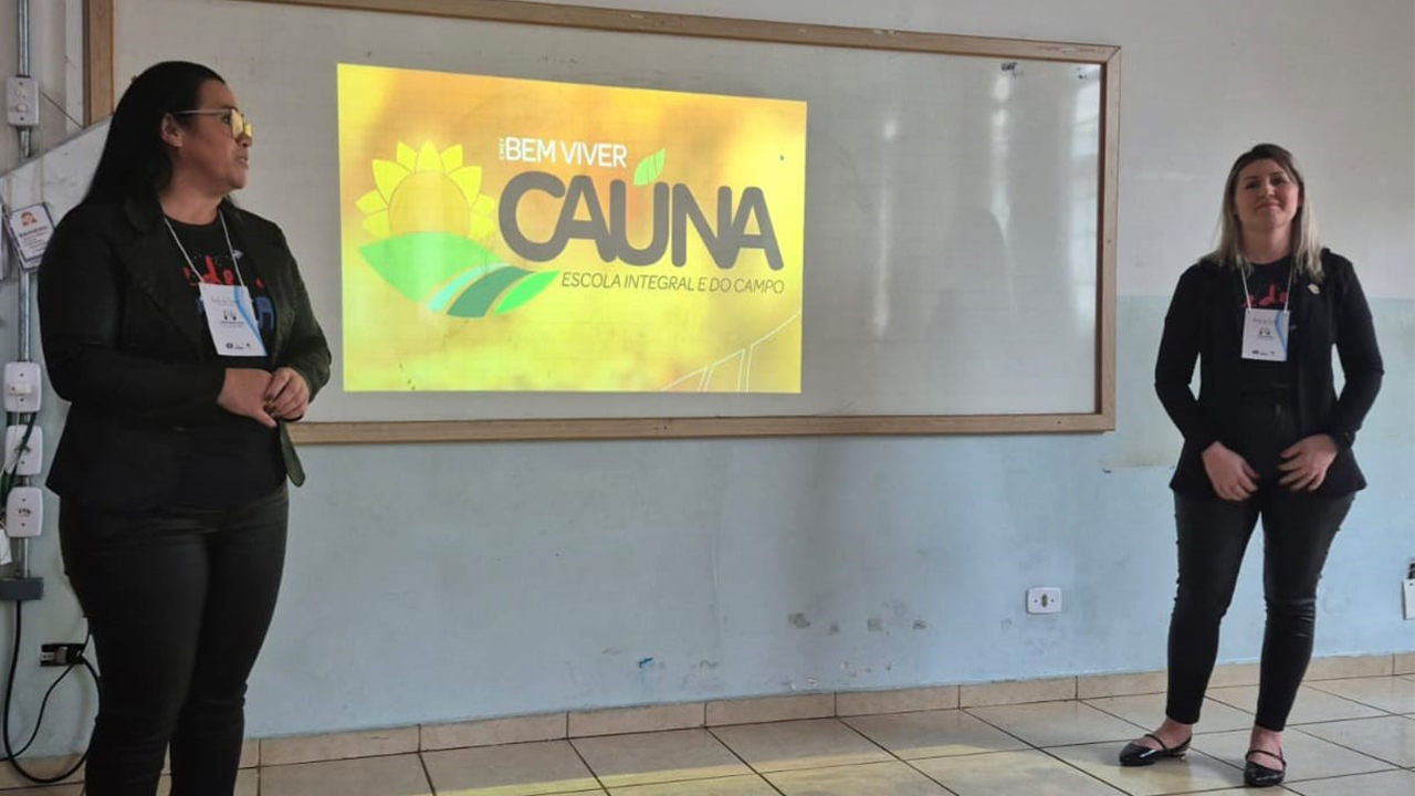 Em uma sala de paredes brancas e piso de cerâmica clara, duas mulheres de roupas escuras e crachás estão diante de uma projeção em uma lousa branca. Na tela, lê-se "Bem Viver Caúna – Escola Integral e do Campo".