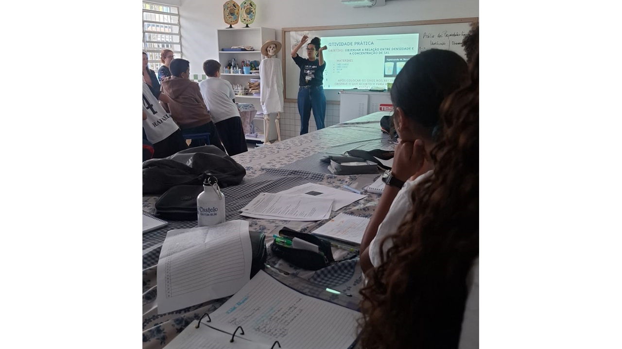 Aula prática em laboratório, com estudantes ao redor de bancadas observando uma mulher à frente, diante de uma projeção. A tela apresenta o texto "Atividade Prática" e conteúdo sobre densidade e concentração de sal. Ao lado dela, há um modelo de esqueleto com chapéu e um pano branco como vestido e uma estante com materiais didáticos. Em primeiro plano, há uma bancada com diversos cadernos, folhas, estojos, garrafas de água e materiais das estudantes.