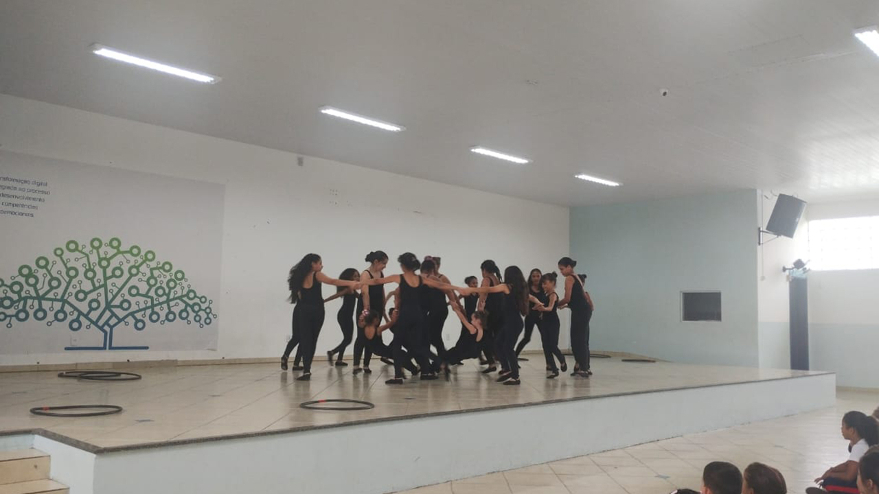 Um grupo multirracial de meninas vestidas de preto realiza uma coreografia, formando uma roda de mãos dadas, com algumas inclinadas para trás. Elas estão sobre um palco elevado em uma sala ampla, com paredes brancas e um painel com a ilustração de uma árvore formada por círculos. À direita, é possível ver uma plateia formada por crianças sentadas no chão de cerâmica clara.