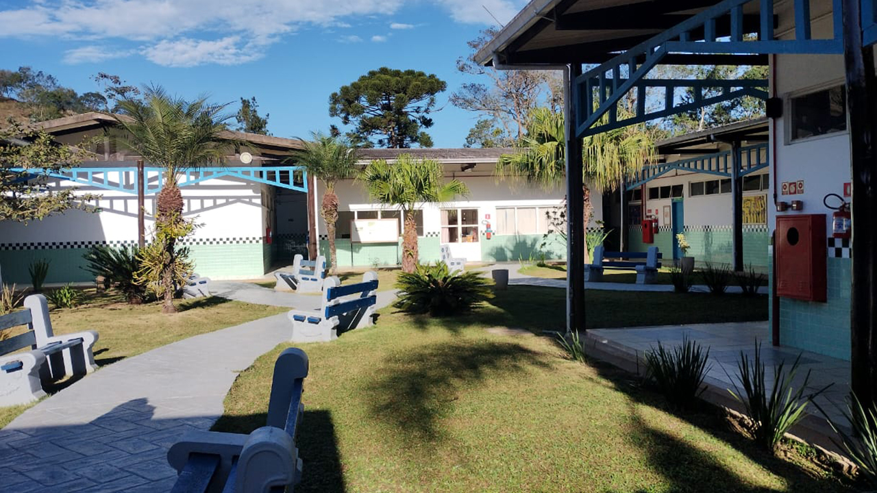 Em um pátio externo, há um caminho sinuoso pavimentado de cinza, contornado por bancos de concreto brancos com assentos azuis dispostos sobre o gramado. O plano de fundo mostra prédios térreos brancos, com esquadrias e estruturas metálicas azuis, faixa de azulejos quadriculados e pequenas palmeiras.