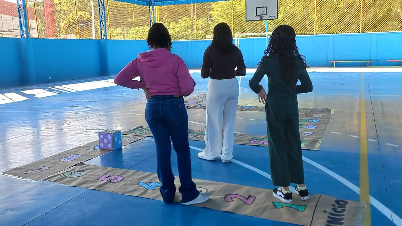 Em uma quadra esportiva, com piso e paredes azuis, três jovens de costas, em trajes casuais, estão em pé, participando de um jogo de tabuleiro em tamanho ampliado. No chão, destaca-se uma trilha de papel com números desenhados e pintados, a palavra "Início" e um dado grande. O fundo exibe uma tabela de basquete e um alambrado amarelo.