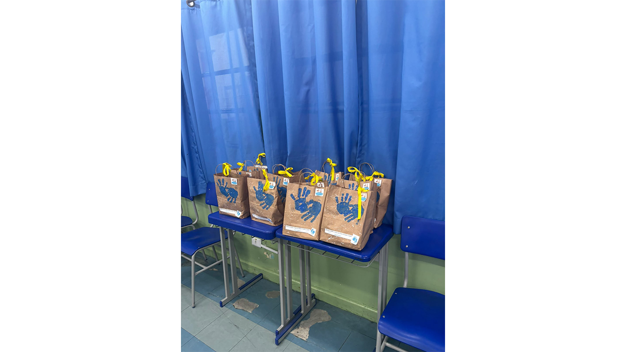 Sacolas de papel pardo, decoradas com marcas de mãos em tinta azul e laços amarelos, estão dispostas sobre duas mesas azuis. O cenário ao fundo é composto por uma longa cortina azul e cadeiras vazias. Também é possível visualizar uma parede verde abaixo das cortinas.