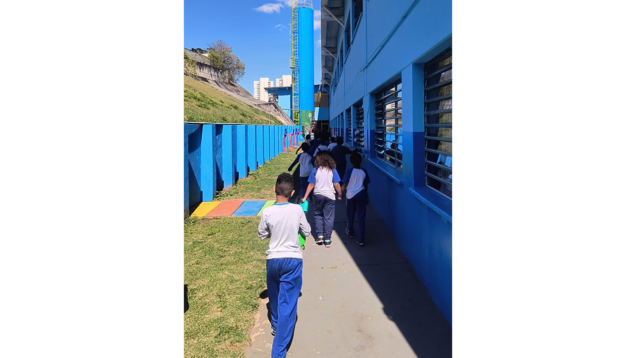 Crianças usando roupas semelhantes, nas cores azul e branco, caminham em fila por um corredor externo. Um caminho de cimento é rodeado por grama. À direita, um prédio azul exibe grandes janelas e, à esquerda, uma mureta azul, mais baixa, com uma encosta gramada ao fundo.