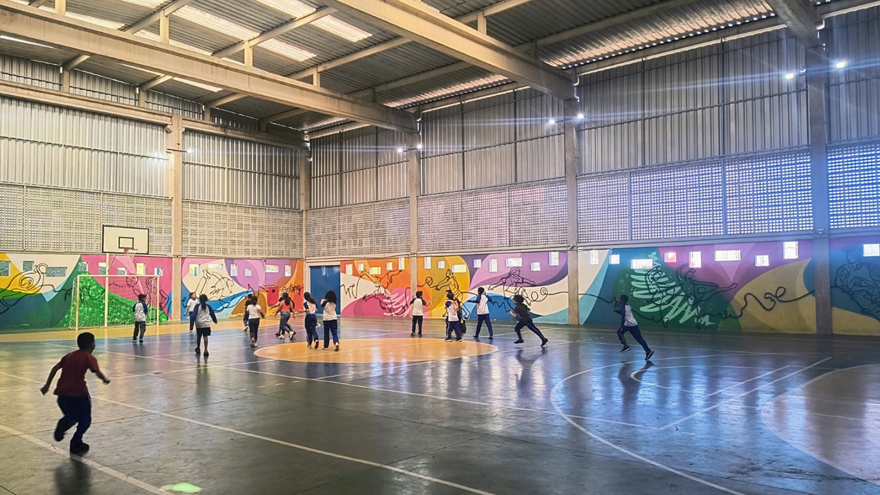 Em uma quadra esportiva escolar, crianças usando camisetas brancas e calças escuras correm dispersas pelo piso liso e brilhante. As paredes exibem um mural multicolorido com desenhos de diferentes práticas esportivas. O fundo revela o teto metálico, paredes com elementos vazados e uma tabela de basquete.