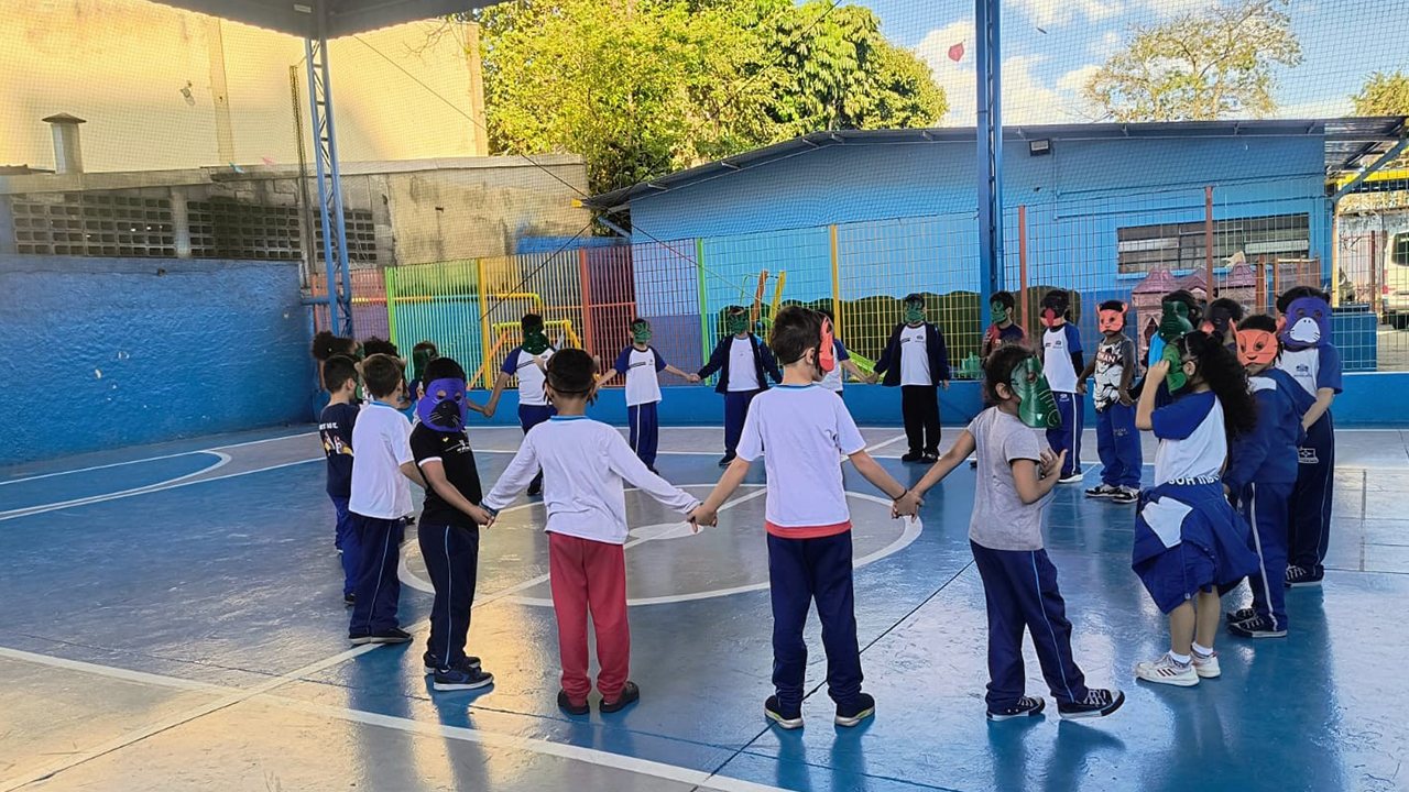 Crianças usando roupas azuis e brancas formam uma roda de mãos dadas. Todas têm o rosto coberto por máscaras coloridas de animais. O piso da quadra é azul, com marcações brancas. O plano de fundo mostra muros azuis, alambrados com grades coloridas, construções e árvores.