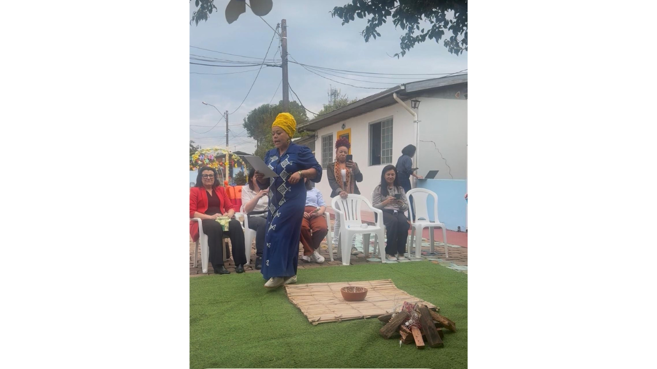 Em uma apresentação externa, uma mulher de vestido azul longo e turbante amarelo lê um papel em pé, observada por pessoas sentadas atrás dela em cadeiras brancas de plástico. Aos seus pés, há um tapete verde que simula grama e, sobre ele, uma esteira de palha, uma tigela e uma fogueira cenográfica. Ao fundo, vê-se uma casa branca e postes de iluminação.