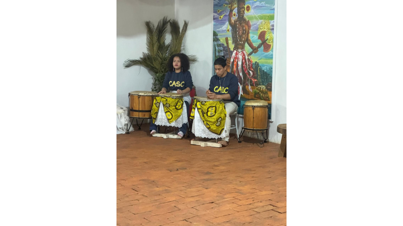 Duas pessoas, vestindo blusas azuis com a sigla "CASC" em amarelo, estão sentadas tocando atabaques de madeira. Os instrumentos estão enfeitados com tecidos estampados em amarelo e rendas brancas. Ao fundo, há um quadro de um homem negro segurando um machado e usando uma saia com fitas brancas e vermelhas. O piso do ambiente é de lajota, e à esquerda da dupla há uma planta.