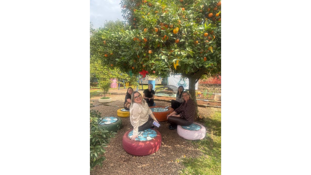 Cinco mulheres, vestindo roupas casuais, estão sentadas em roda. O grupo acomoda-se sobre pneus coloridos, reaproveitados como bancos, com tampos de mosaico azul. O cenário é sombreado por uma árvore cheia de frutos na cor laranja. Ao redor, é possível ver canteiros, chão de terra e de pedras.
