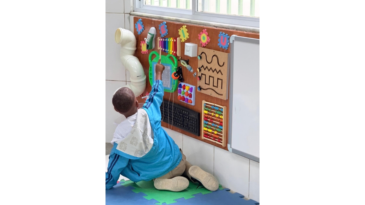 Ambiente escolar com painel sensorial fixado na parede. Uma criança, sentada de costas sobre placas de EVA verdes e azuis, toca um xilofone colorido com uma baqueta. O painel reúne letras de EVA, ábaco, teclado de computador, um pedaço de cano branco, cadarço, formas e outros objetos táteis. Ao fundo, há uma janela e uma parede revestida.