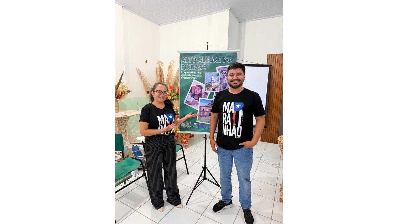 Em uma sala interna de paredes, teto e piso branco, uma mulher e um homem, ambos vestindo camisetas pretas estampadas com o nome "MARANHÃO" e uma estrela branca num fundo azul, posam diante de um banner verde com fotos e o título "Alto Alegre do Pindaré". Ao redor, observam-se cadeiras, mesas e elementos decorativos de palha.