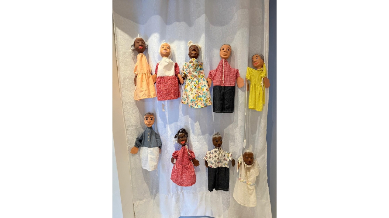 Exposição de nove bonecos artesanais pendurados sobre um tecido branco. As figuras representam homens e mulheres com diferentes tons de pele, cabelos grisalhos em sua maioria e roupas variadas em cores e estampas.