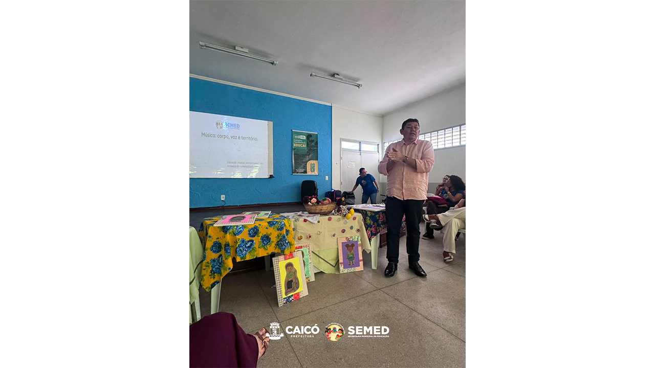 Sala de apresentação com um homem em pé, falando ao público, diante de mesas com bonecas, tecidos e toalhas amarelas com flores azuis e vermelhas e materiais pedagógicos. Em uma parede azul, uma tela de projeção tem o texto: "Música: corpo, voz e território".