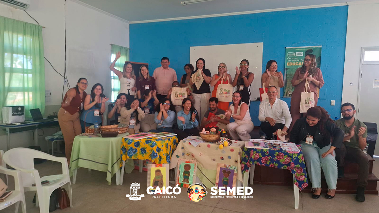 Grupo multirracial de 20 pessoas posa para foto de forma espontânea, batendo palmas, sorrindo e brincando entre si. Algumas estão em pé, outras agachadas, e várias seguram sacolas de estilo eco bag com a inscrição “Eco Sala”. O grupo está em cima de um palco marrom escuro, à frente da sala. Perto deles, no chão, há mesas cobertas por tecidos estampados que exibem bonecas de pano, livros e materiais pedagógicos. Ao fundo, a parede é azul e conta com uma lousa branca.