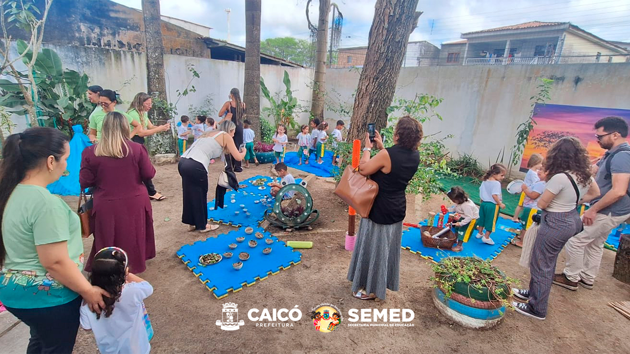 Área externa arborizada de terra batida com crianças e adultos participando de atividades pedagógicas distribuídas pelo espaço. As crianças exploram objetos e materiais naturais organizados sobre placas azuis de EVA no chão, enquanto os adultos acompanham e registram a atividade. Ao fundo, há árvores, plantas, um muro e um painel com imagem de paisagem.