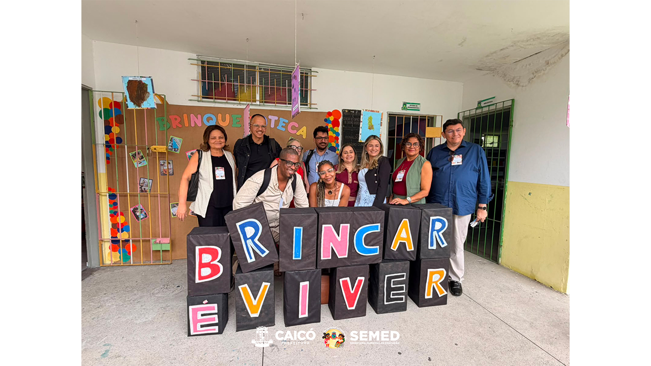 Grupo multirracial formado por dez adultos posa para foto em uma área coberta. À frente, grandes cubos pretos formam a frase “BRINCAR É VIVER”, com letras coloridas. Ao fundo, há um painel de papel pardo com a palavra “BRINQUEDOTECA” em letras coloridas, fotografias, balões e portas gradeadas nas cores verde e amarela. O chão é cinza e as paredes são brancas e amarelas.