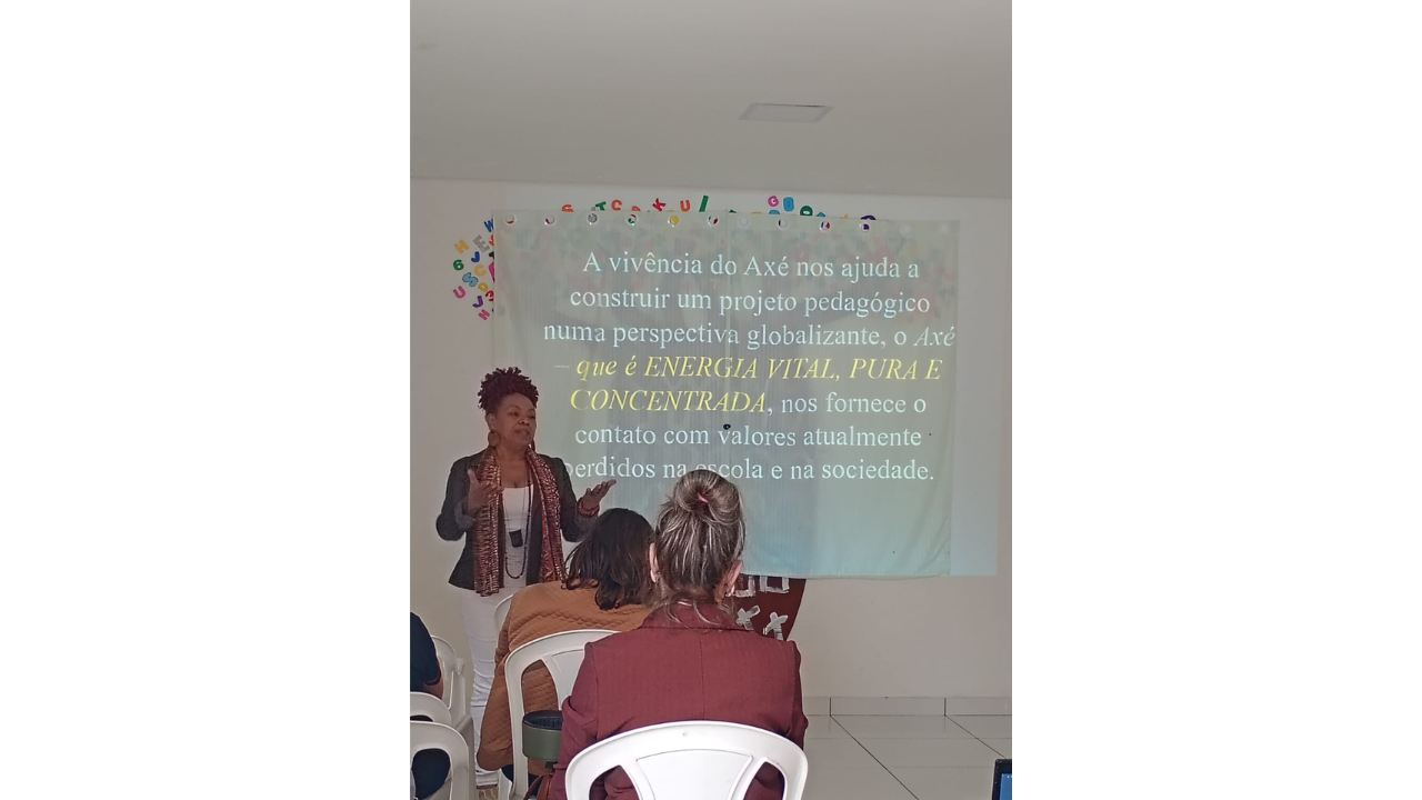 Uma mulher em pé, de casaco escuro e echarpe estampada, gesticula enquanto fala para uma plateia sentada de costas, em cadeiras brancas de plástico. O grupo está em uma sala de apresentações, onde há uma projeção na parede branca, com o texto "A vivência do Axé nos ajuda a construir um projeto pedagógico numa perspectiva globalizante, o Azé que é ENERGIA VITAL, PURA E CONCENTRADA, nos fornece o contato com valores atualmente perdidos na escola e na sociedade."
