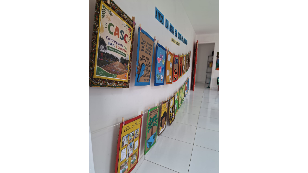 Em um corredor claro de chão e paredes brancas, uma exposição de trabalhos manuais está pendurada em dois varais na parede. As produções têm molduras coloridas e, na primeira delas, é possível ler "CASC", em letras verdes e laranjas. Ao fundo, uma porta aberta.