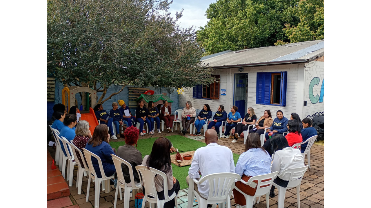 Reunião de cerca de vinte e cinco pessoas multirraciais em um pátio. Vestindo trajes casuais, estão sentadas em roda, em cadeiras plásticas. No centro, sobre a grama, há uma esteira de palha, uma tigela e uma fogueira cenográfica. O fundo mostra uma árvore, piso de tijolos e uma casa branca com janelas azuis. O ambiente também conta com brinquedos infantis de plástico, como um escorregador laranja e uma casinha de teto verde.