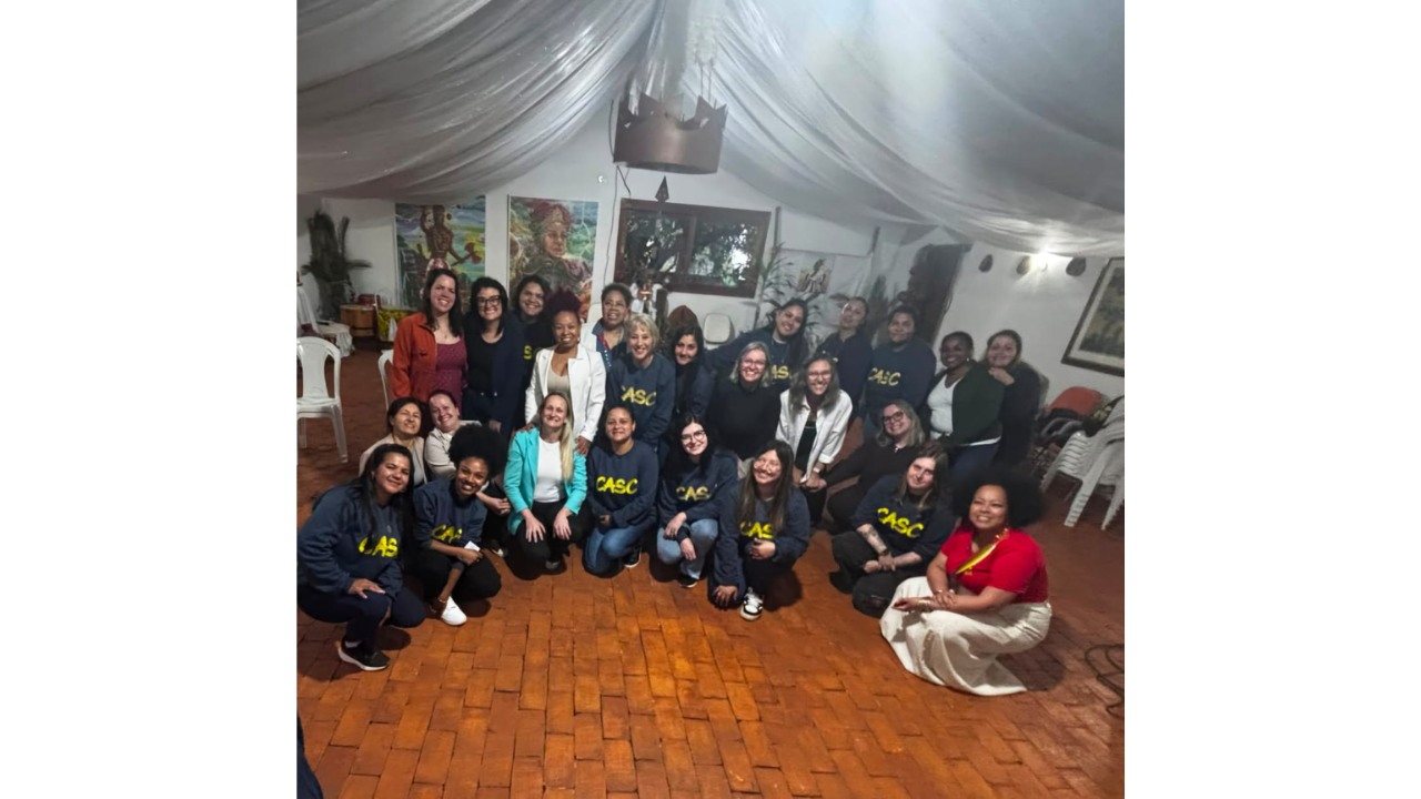 Um grupo multirracial de cerca de 25 mulheres está reunido, com pessoas em pé e agachadas, sorrindo. A maioria veste moletons azuis com "CASC" escrito em amarelo. O espaço tem piso de tijolos vermelhos, paredes brancas com quadros e teto forrado com panos brancos drapeados.