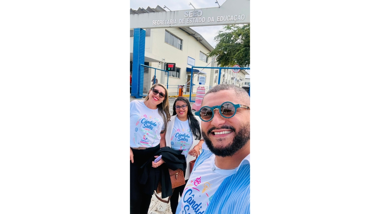 Grupo multiracial composto por duas mulheres e um homem, usando camisetas brancas com inscrições em azul “Cândido Sales vai à Rede de Trocas 2026”, posa para uma selfie. Ao fundo, aparecem o portão azul de ferro e uma fachada com um letreiro cinza: "SEED – Secretaria de Estado da Educação". Ao fundo, é possível avistar uma árvore e uma construção baixa, de dois andares, em amarelo claro.