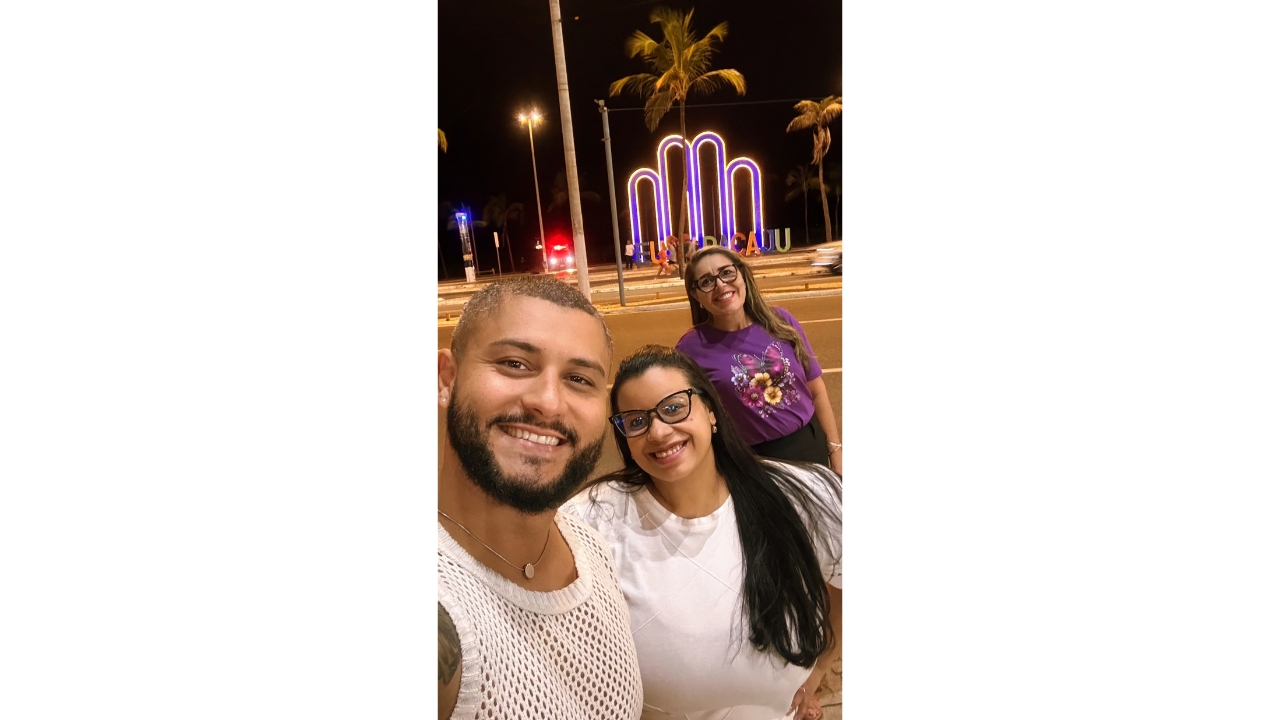 Área urbana à noite, onde um grupo multirracial de três adultos faz uma selfie e sorri para a câmera. Em primeiro plano, há um homem e duas mulheres. Atrás do grupo, uma estrutura iluminada na cor roxo neon forma um símbolo e o nome “Aracaju” está aos apoiado no chão, entre palmeiras e iluminação pública.