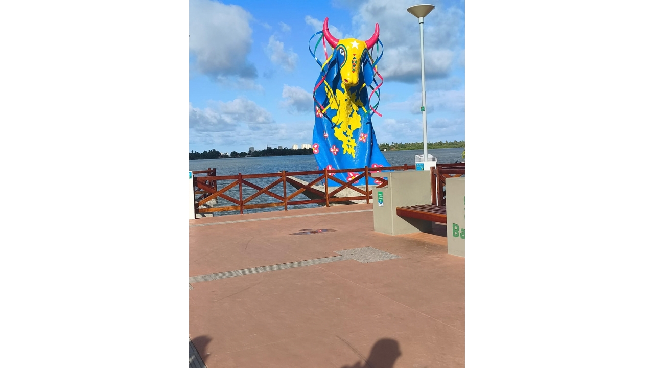 Área externa à beira d’água, com uma grande escultura de um boi. A peça é azul, com cabeça e peito amarelos, chifres vermelhos, flores e fitas coloridas. Ao redor, há guarda-corpo de madeira escura, um banco, poste de luz e, ao fundo, água e vegetação.