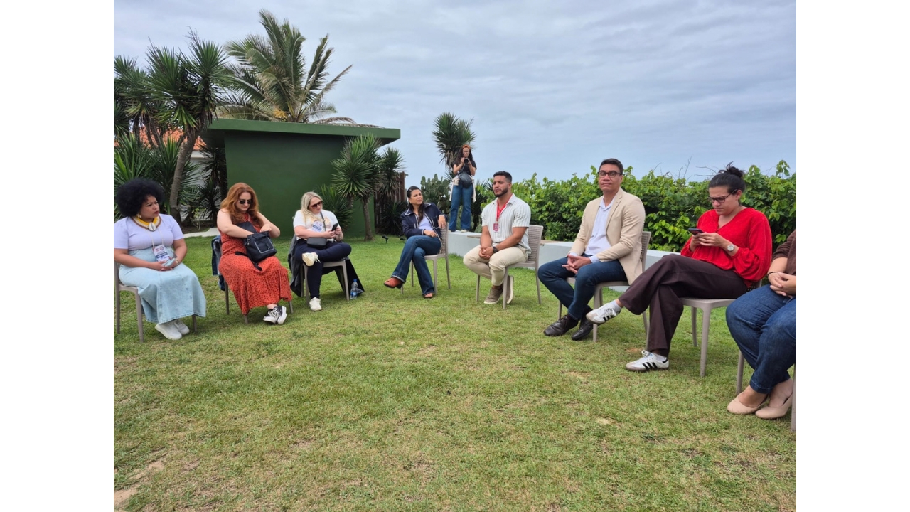 Atividade ao ar livre em área gramada, com um grupo multirracial de adultos sentados em círculo. As(os) participantes usam roupas casuais e alguns crachás; uma pessoa permanece em pé ao fundo, tirando fotos. O espaço é cercado por vegetação, palmeiras e arbustos e um pequena construção verde.