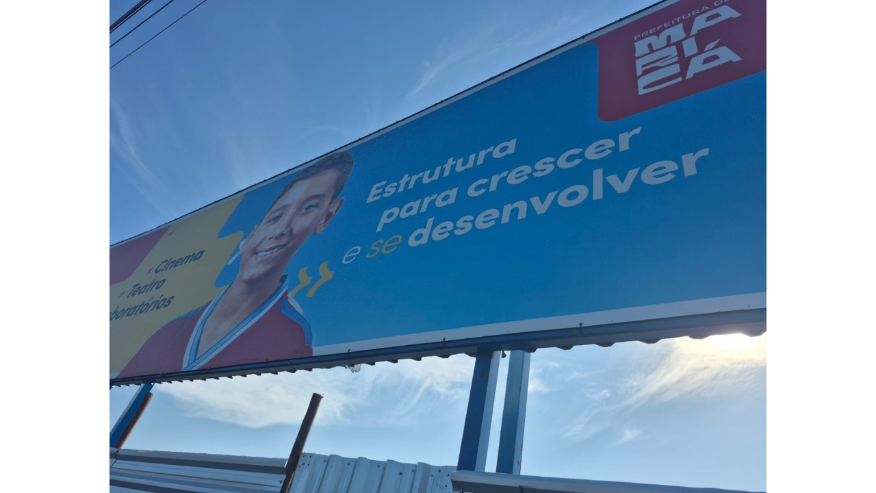 Área externa com grande painel institucional (outdoor), com a imagem de uma criança e a frase “Estrutura para crescer e se desenvolver” em letras brancas e um fundo azul, além da identificação da Prefeitura de Maricá em um fundo vermelho.
