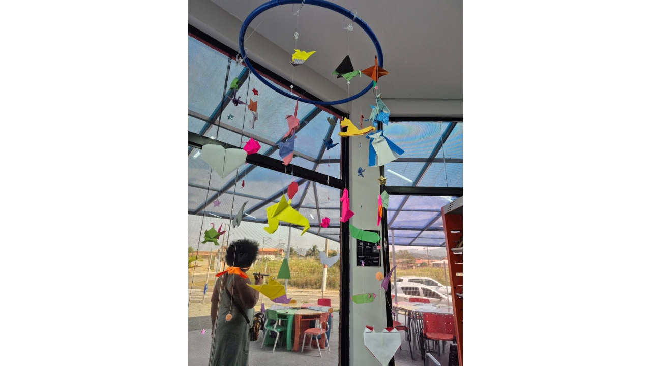 Espaço interno decorado com um móbile de origami suspenso em um aro azul. As dobraduras têm diferentes formas e cores. Ao fundo, grandes portas e janelas de vidro, mesas e cadeiras coloridas e uma pessoa em pé próxima à área externa.