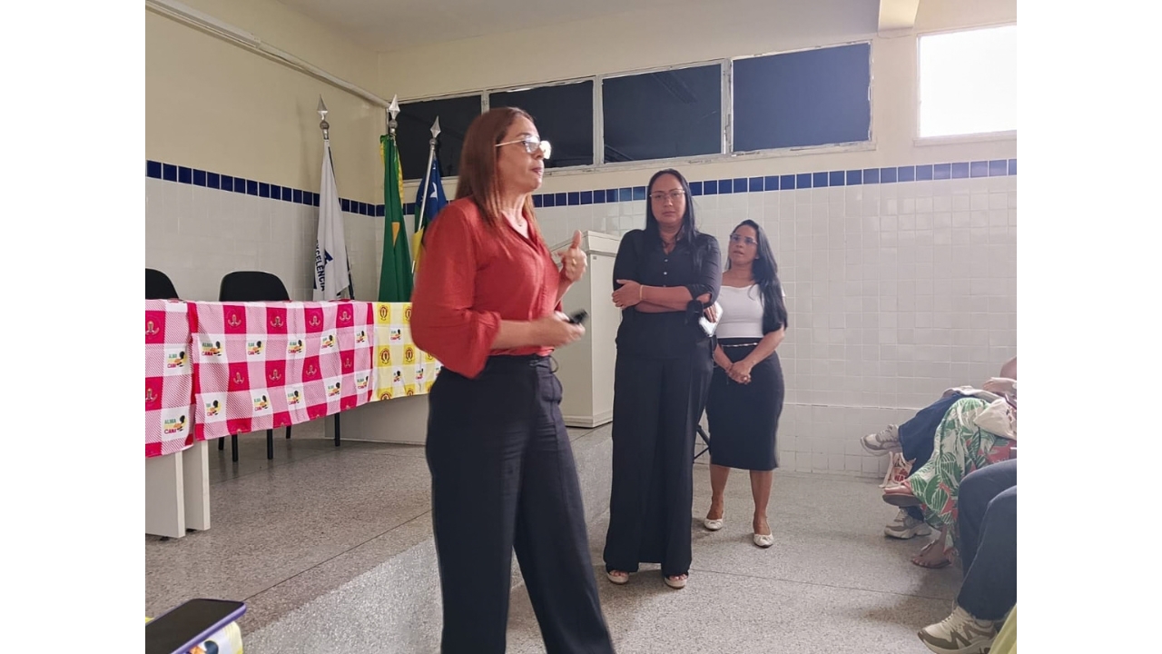 Sala de apresentação com público sentado à direita, com pernas parcialmente visíveis. Uma mulher de blusa vermelha e calça preta fala em pé, enquanto duas mulheres - vestindo calça e blusa preta e saia preta e blusa branca - permanecem ao fundo, observando. À esquerda, há uma mesa longa decorada com duas toalhas xadrez rosa e amarela e bandeiras ao fundo.