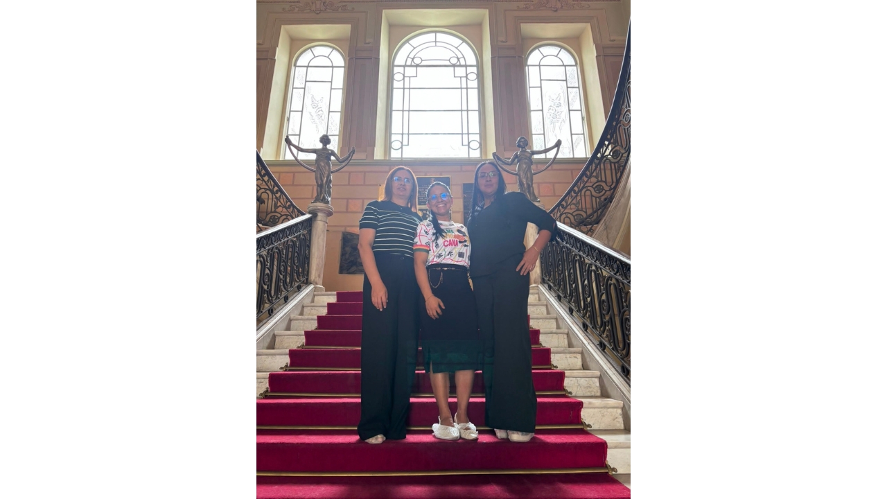 Ambiente com escadaria de mármore coberta por um tapete vermelho centralizado. Um grupo multirracial de três mulheres posa lado a lado nos degraus; duas vestem roupas pretas e uma usa camiseta estampada. Ao fundo, há três grandes janelas arqueadas, corrimãos ornamentados e duas esculturas metálicas, uma de cada lado da escadaria.