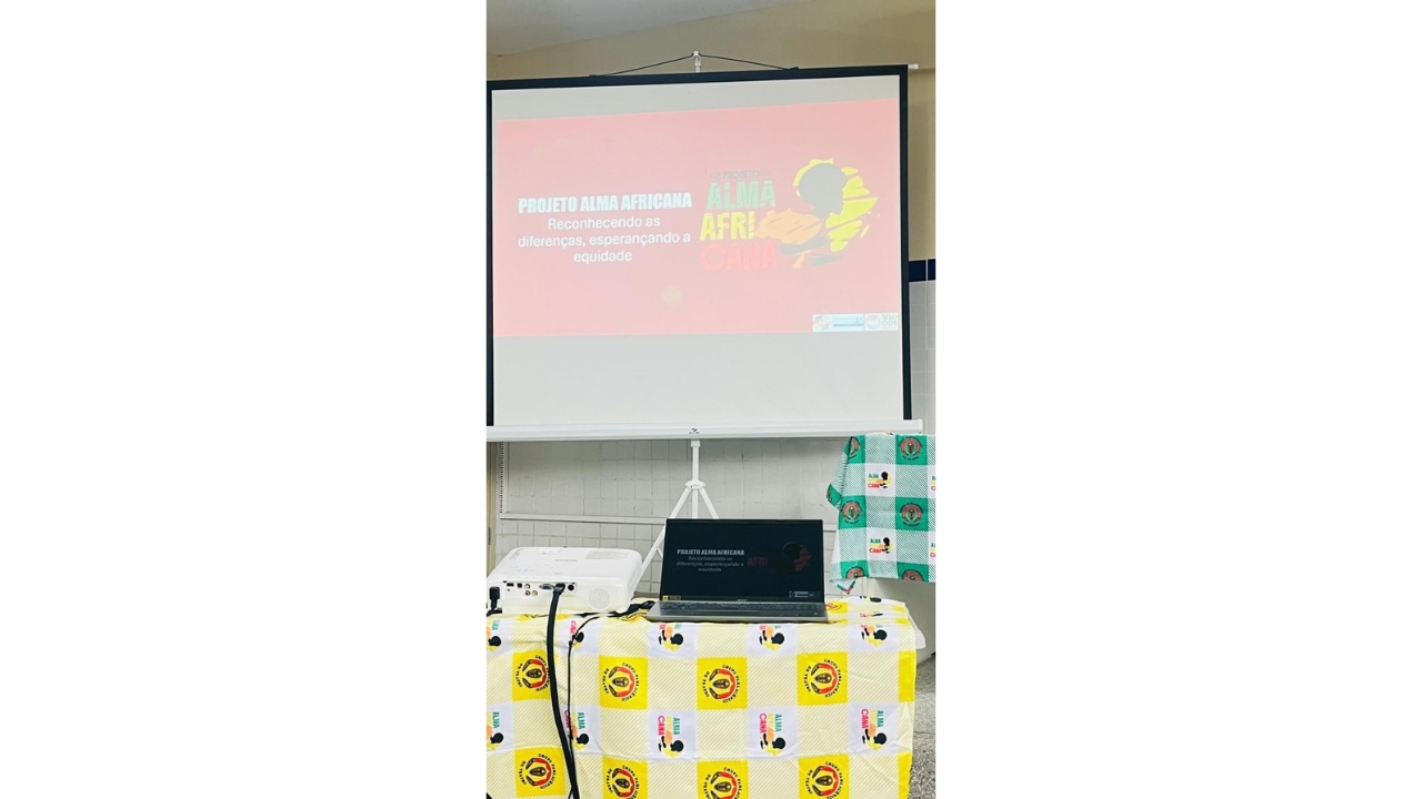 Espaço de apresentação com tela de projeção e equipamentos sobre uma mesa. Na tela, lê-se “Projeto Alma Africana – Reconhecendo as diferenças, esperançando a equidade”, em um fundo vermelho e um logo nas cores verde, amarelo e vermelho, com o contorno do continente africano. Em primeiro plano, em cima de uma mesa com uma toalha xadrez branca e amarela com estampa, há um notebook aberto e um projetor branco ao lado.